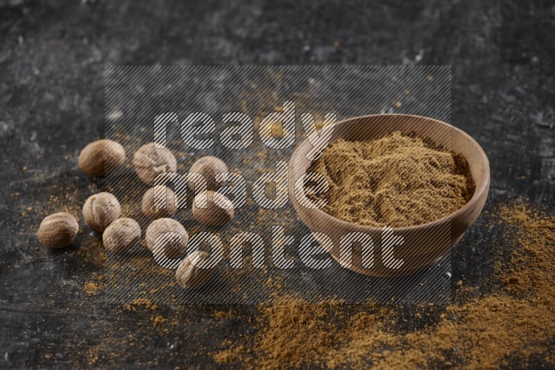 A wooden bowl full of nutmeg powder with whole seeds and sprinkled powder beside it on a textured black flooring