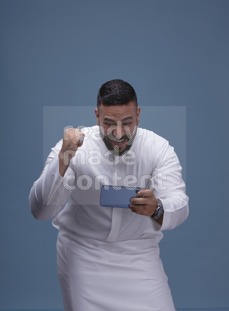 A man Playing a Game on his Smartphone  on Blue Background wearing Saudi Thob