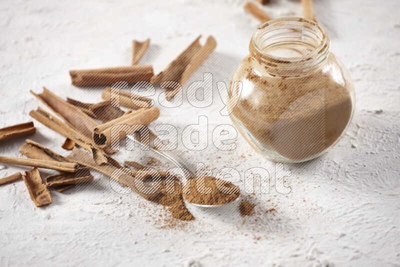 Herbal glass jar full cinnamon powder and a metal spoon surrounded by cinnamon sticks on a white background