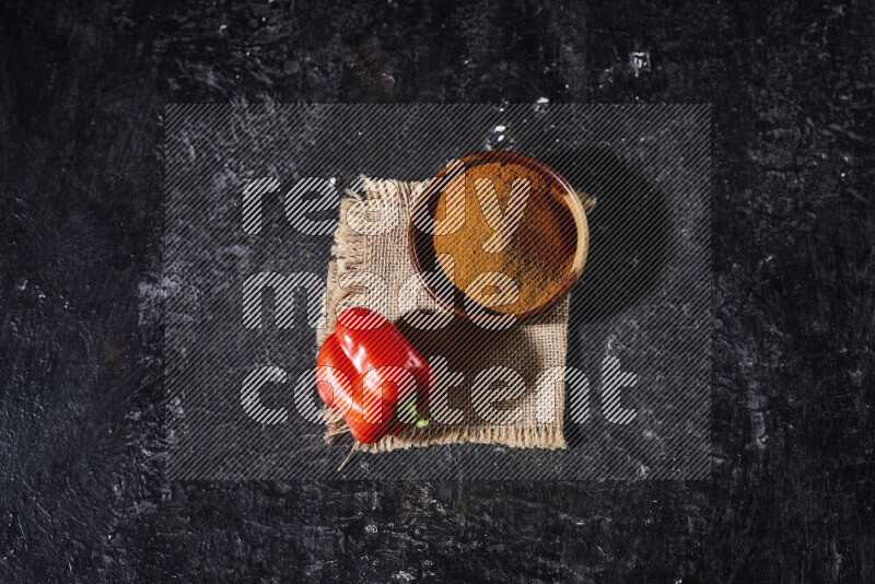 A wooden bowl full of ground paprika powder with a red bell pepper on a burlap fabric on black background