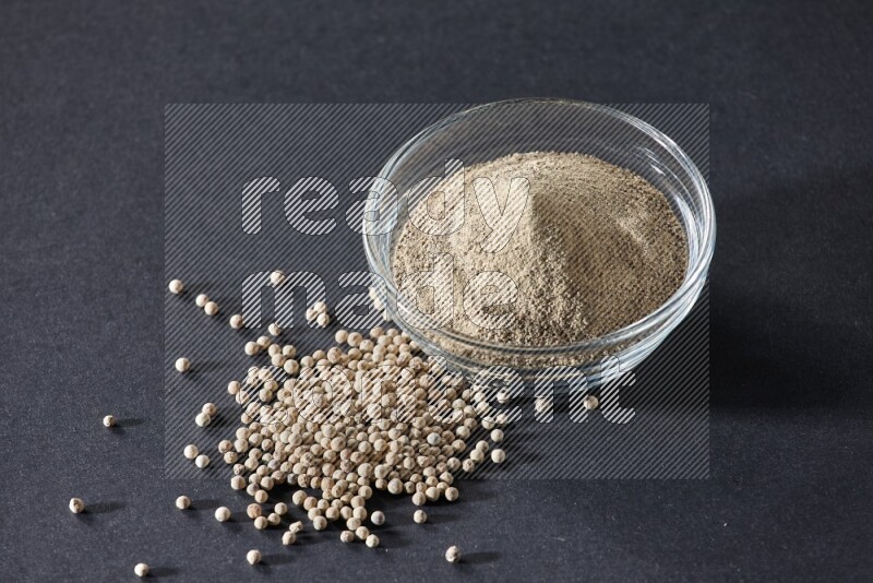 A glass bowl full of white pepper powder with white pepper beads on black flooring