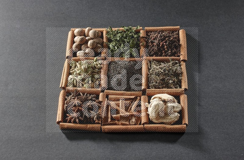 9 squares of cinnamon sticks full of tea in the middle surrounded by nutmeg, dried mint, cloves, dried basil, dried ginger, cinnamon, star anise and cardamom on black flooring