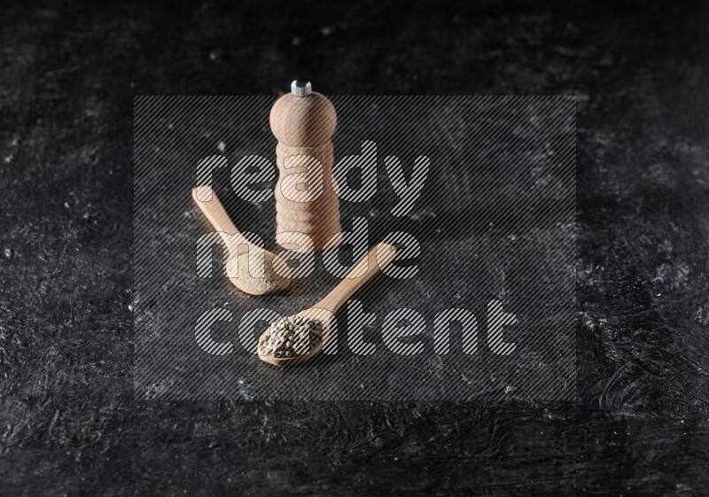 2 wooden spoons one full of white pepper powder and the other with pepper beads and a wooden pepper mill on textured black flooring