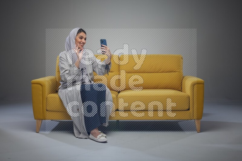 A Saudi woman wearing a light gray Abaya and head scarf sitting on a yellow sofa and making a video call eye level on a grey background