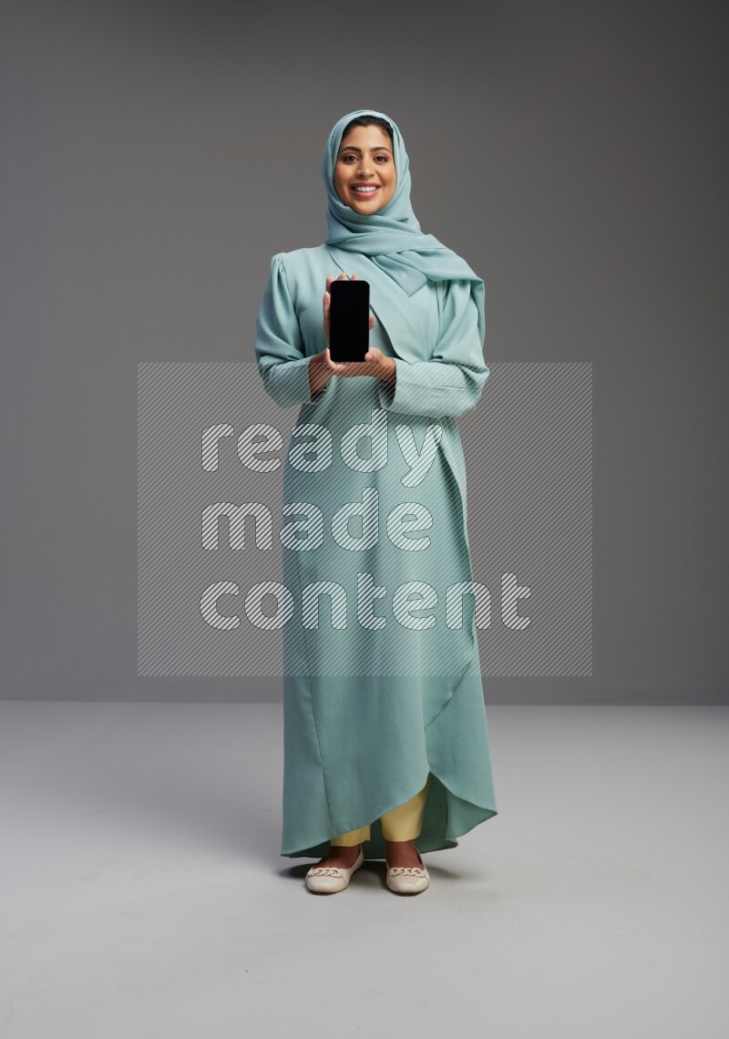 Saudi Woman wearing Abaya standing showing phone to camera on Gray background