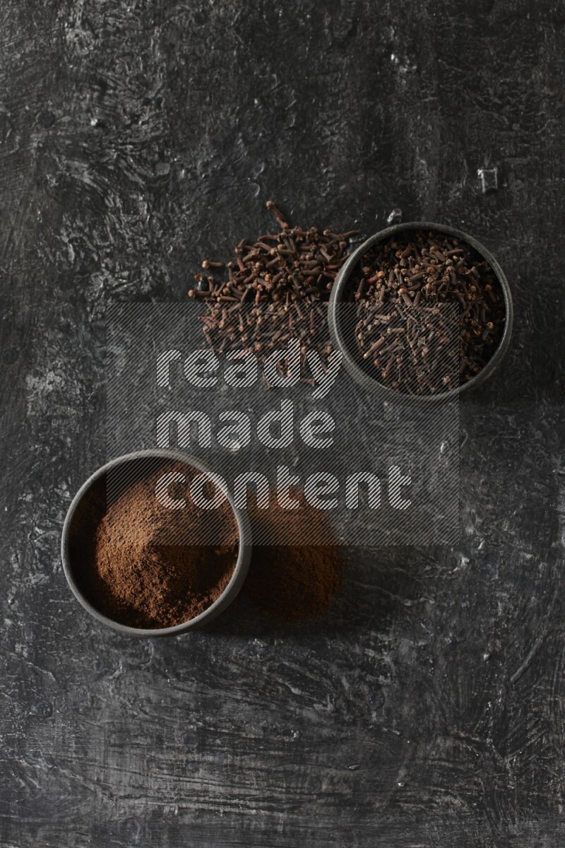 2 Black pottery bowls full of cloves and the other full of cloves powder on textured black flooring