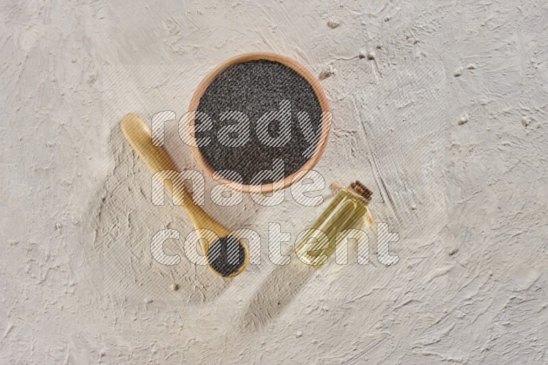 A wooden bowl and spoon full of black seeds with a bottle of black seeds oil on a textured white flooring