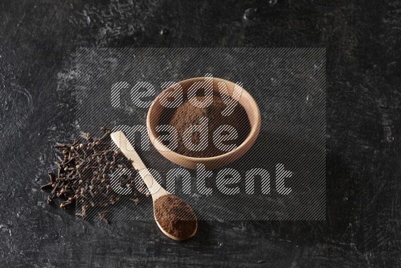 A wooden bowl and a wooden spoon full of cloves powder with spreaded cloves on a textured black flooring