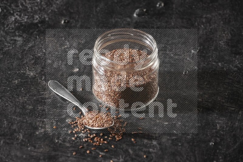 A glass jar full of flaxseeds with a metal spoon full of the seeds on a textured black flooring