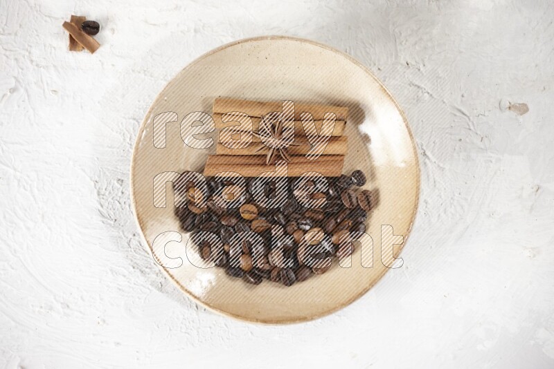 Beige plate full of coffee beans, cinnamon sticks and star anise with a coffee grinder, coffee beans, cinnamon pieces and cardamom next of it on white background