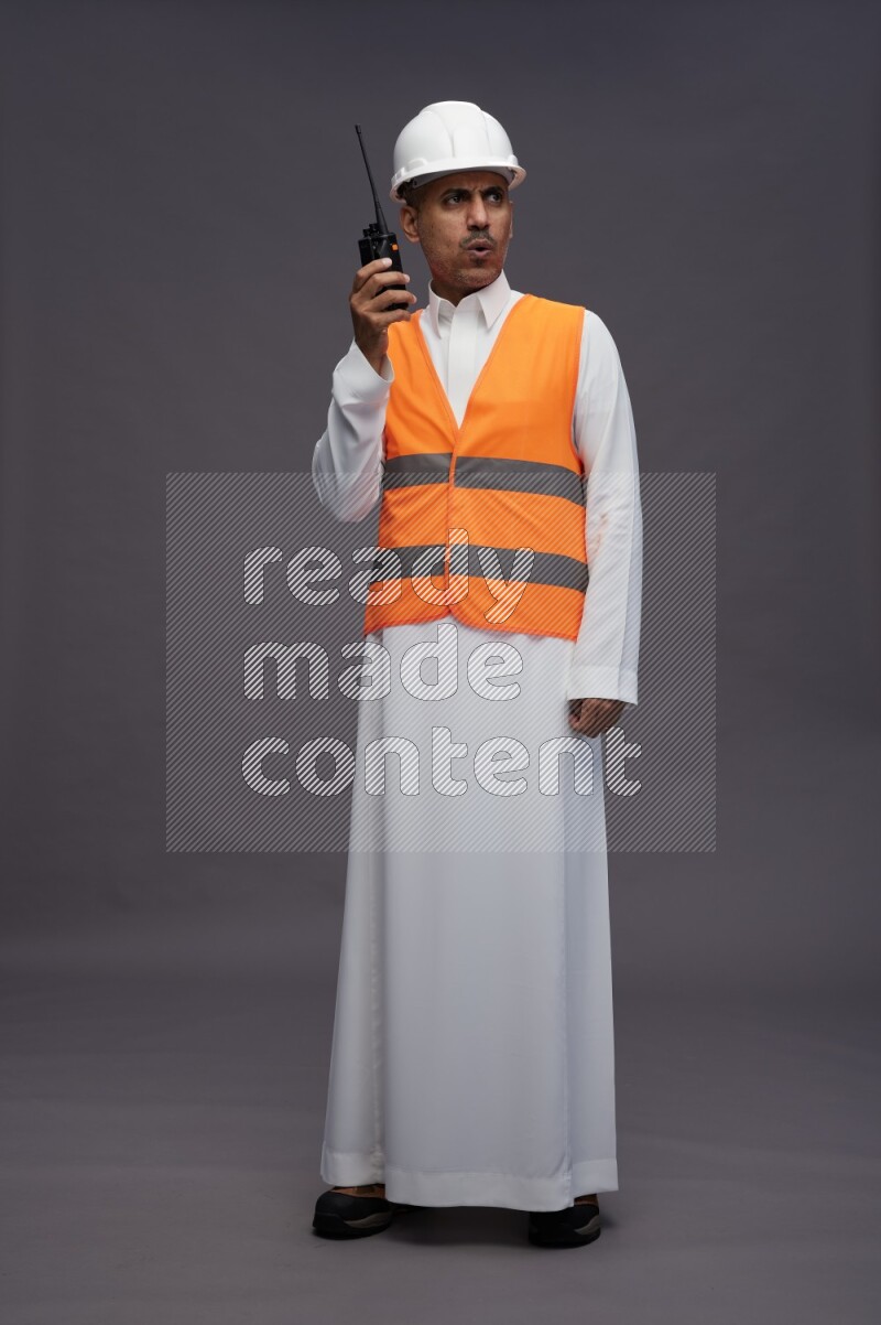 Saudi man wearing thob with engineer vest standing holding walkie-talkie on gray background