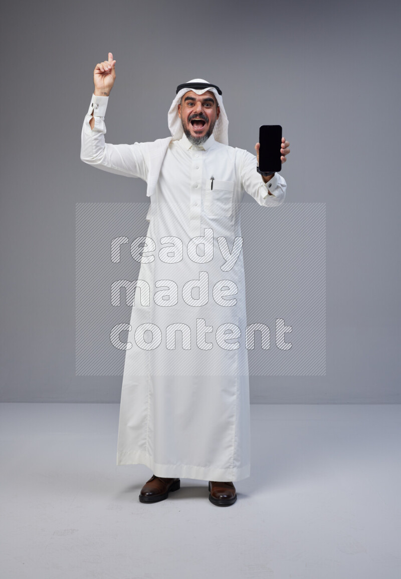 Saudi man Wearing Thob and white Shomag standing showing phone to camera on Gray background