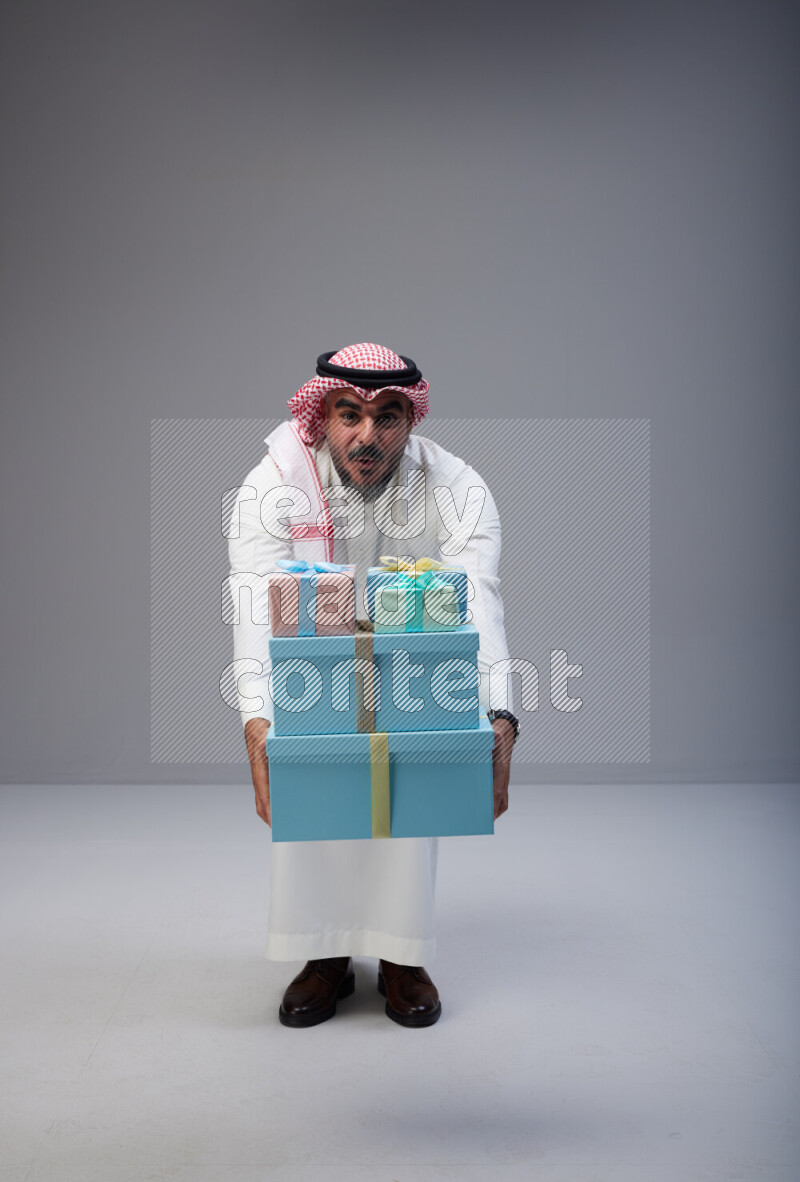 Saudi man Wearing Thob and red Shomag standing holding gift box on Gray background