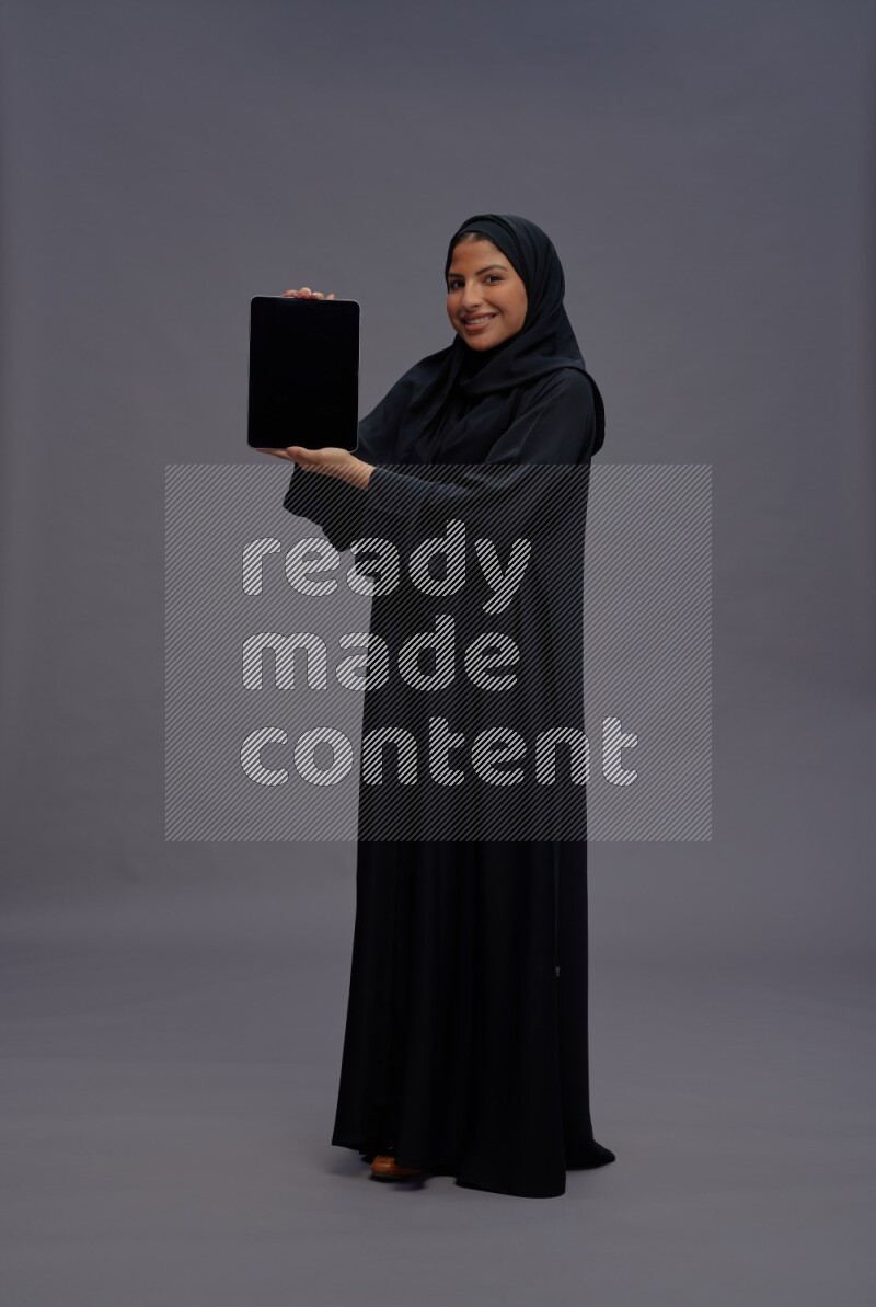 Saudi woman wearing Abaya standing showing tablet to camera on gray background