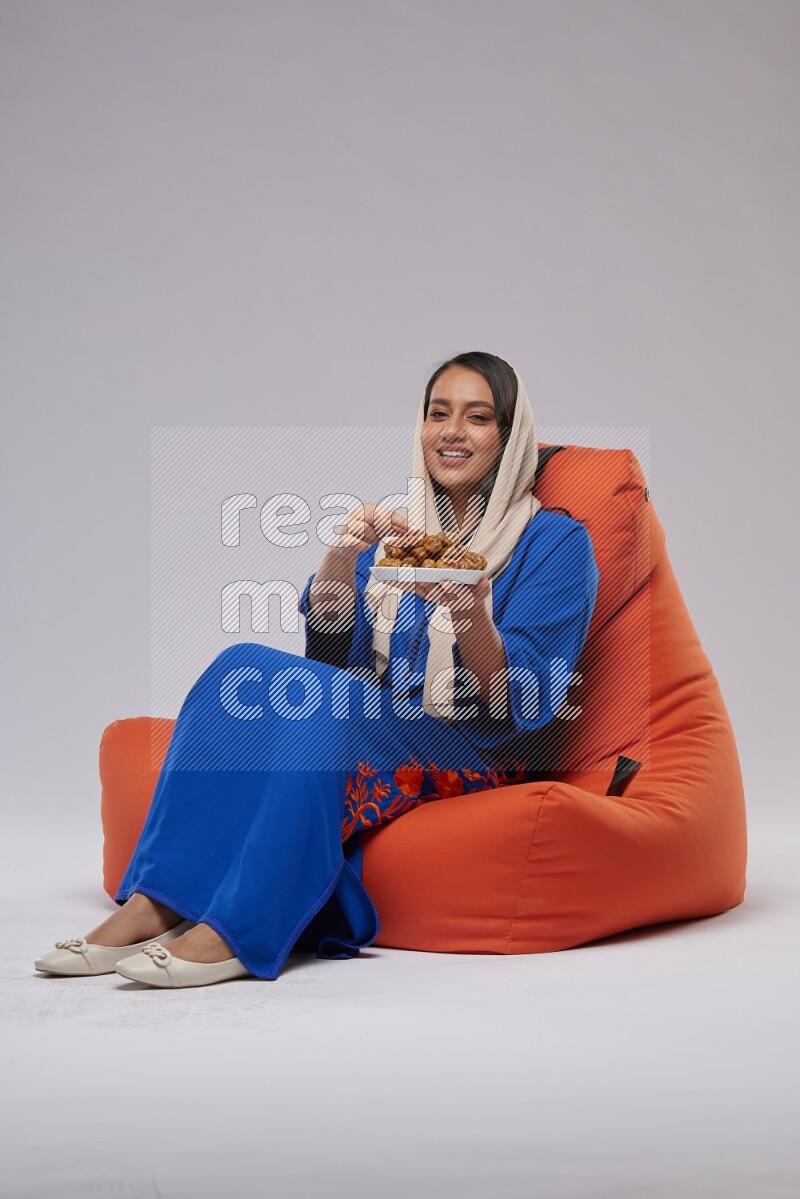 A Woman sitting on an orange beanbag wearing Jalabeya holding a plate of dates
