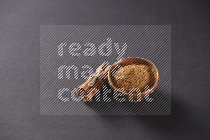 Cinnamon sticks stacked and bounded beside a wooden bowl full of cinnamon powder and a wooden spoon full of powder on black background