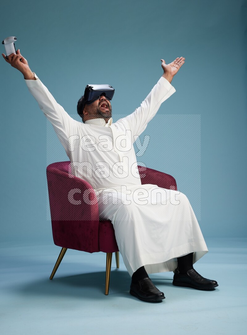 Saudi Man without shimag sitting on chair  gaming with VR and joystick on blue background