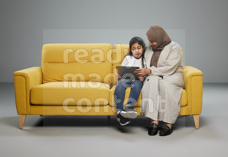 A girl with her mother on yellow sofa and watching on iPad on gray background