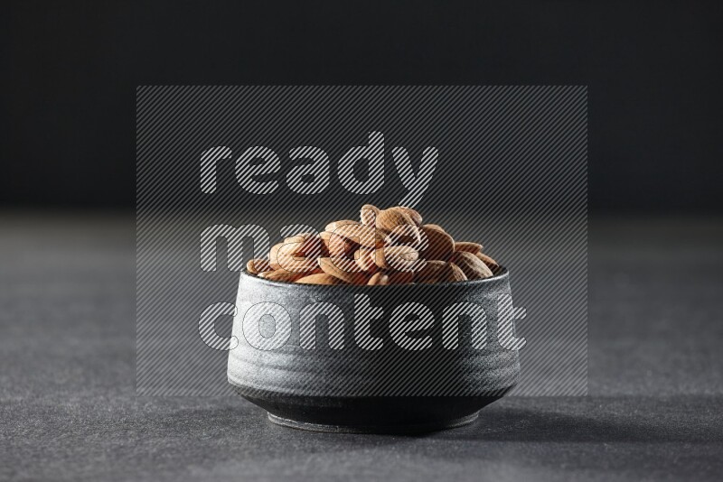 A black pottery bowl full of peeled almonds on a black background in different angles