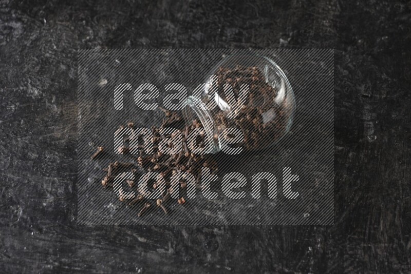 A flipped glass spice jar full of cloves and cloves came out of it on textured black flooring