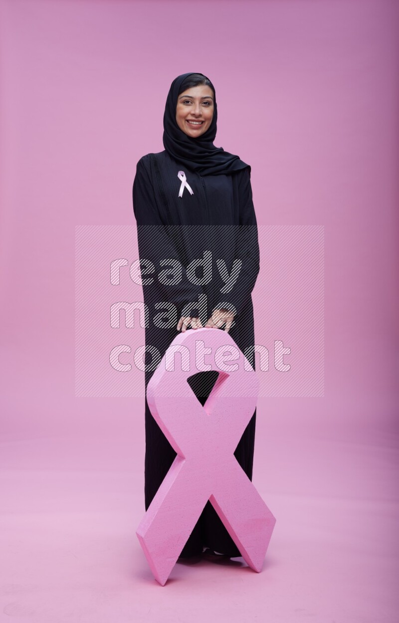 Saudi woman wearing pink ribbon on abaya standing holding awareness ribbon on plain pink background