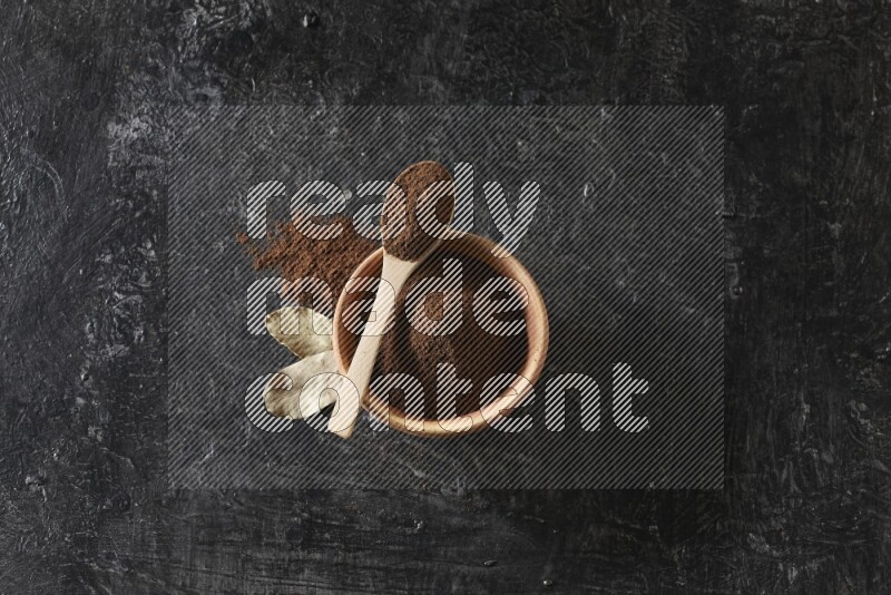 A wooden bowl and a wooden spoon full of cloves powder with laurel leaves on a textured black flooring