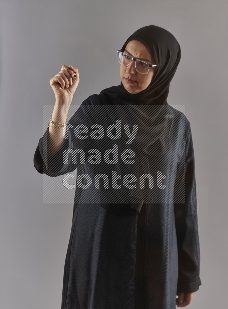 Woman in black abaya, hijab and glasses holding a marker pen to write on a transparent board with different reactions and expressions on grey background