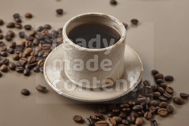 A beige pottery cup of coffee surrounded by roasted coffee beans on beige background