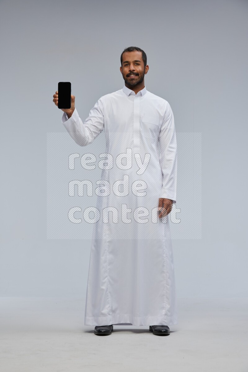 Saudi man wearing Thob standing showing phone to camera on Gray background