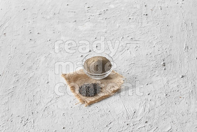 A glass bowl full of black pepper powder and black pepper beads on burlap fabric on textured white flooring