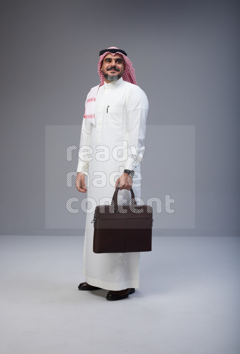 Saudi man Wearing Thob and red Shomag standing holding bag on Gray background