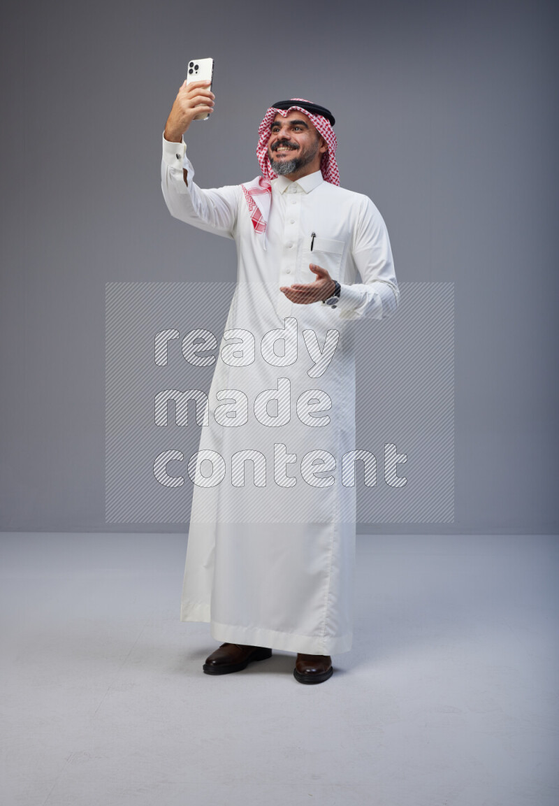 Saudi man Wearing Thob and red Shomag standing taking selfie on Gray background