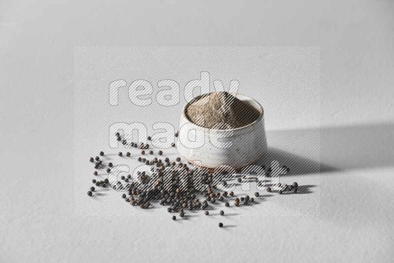 A white ceramic bowl full of black pepper powder and black pepper beads spread on white flooring