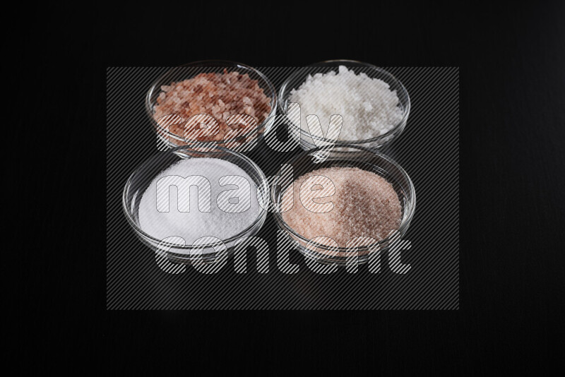 4 glass bowls filled with table salt, coarse sea salt, fine himalayan salt and coarse himalayan salt on black background