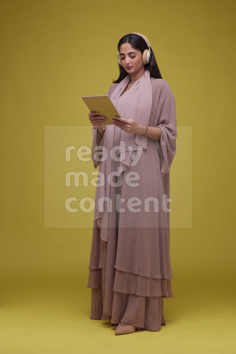 A woman Holding an Ipad with Headphone on a Yellow Background wearing Brown Abaya