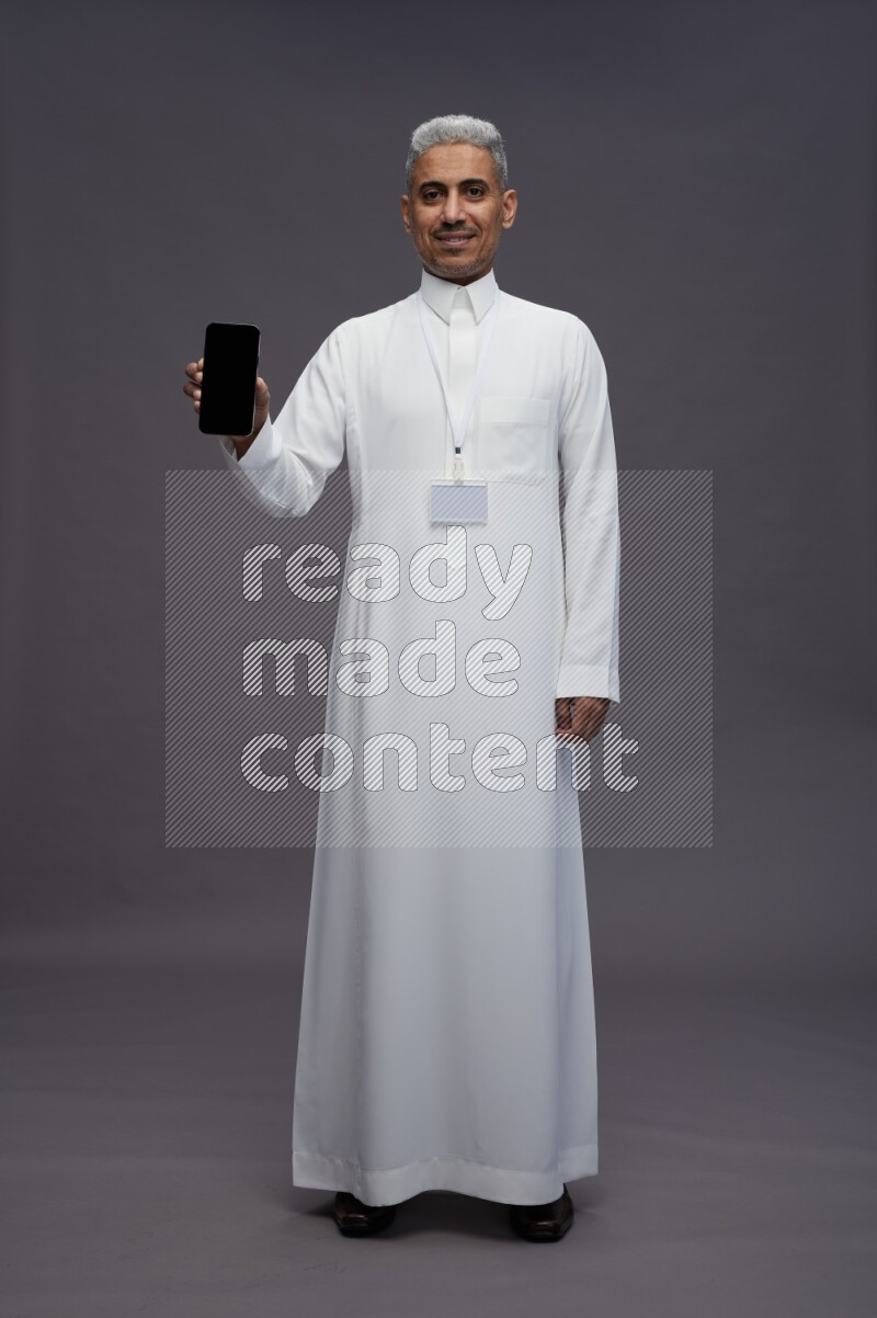Saudi man wearing thob with neck strap employee badge standing showing phone to camera on gray background