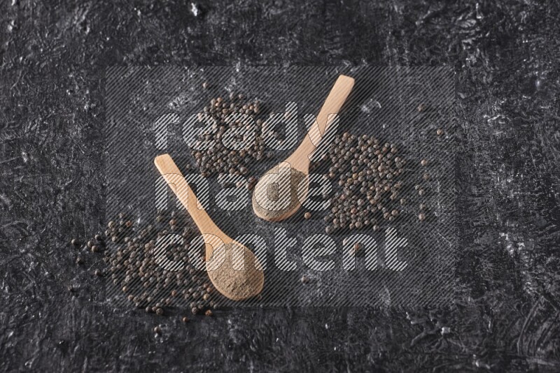 2 wooden spoons full of black pepper powder and black pepper beads spread on a textured black flooring