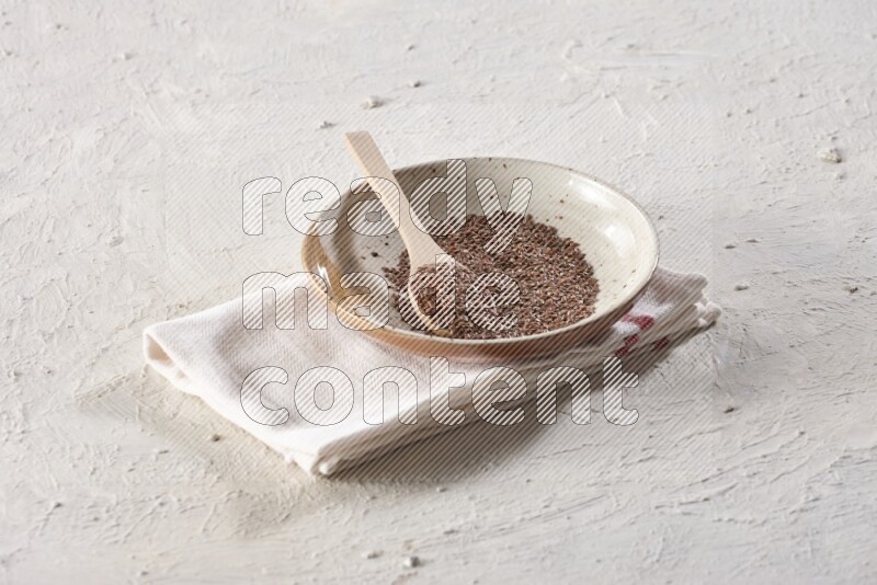 A multicolored pottery plate full of flax seeds with a wooden spoon full of the seeds on a napkin on a textured white flooring