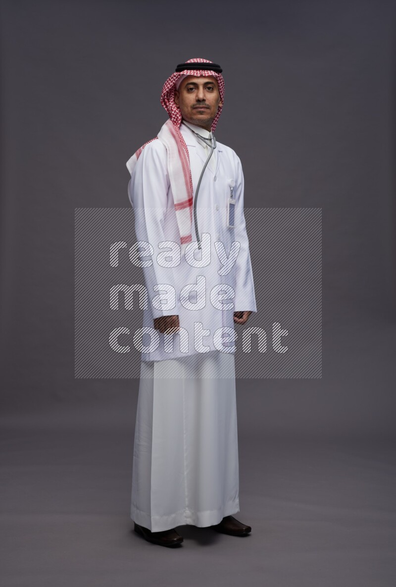 Saudi man wearing thob with lab coat and shomag with pocket employee badge with stethoscope standing interacting with the camera on gray background