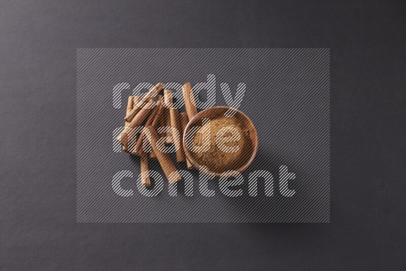 Cinnamon sticks stacked beside a wooden bowl full of cinnamon powder on black background