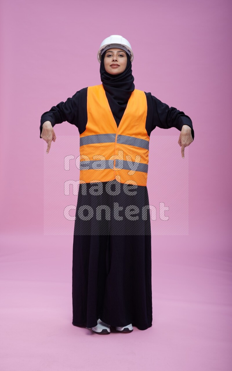 Saudi woman wearing Abaya with engineer vest and helmet standing interacting with the camera on pink background