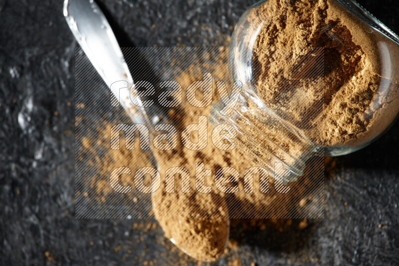 A flipped glass spice jar and a metal spoon full of allspice powder and powder spilled out of it on a textured black flooring