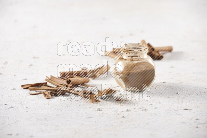 Herbal glass jar full cinnamon powder and a metal spoon surrounded by cinnamon sticks on a white background