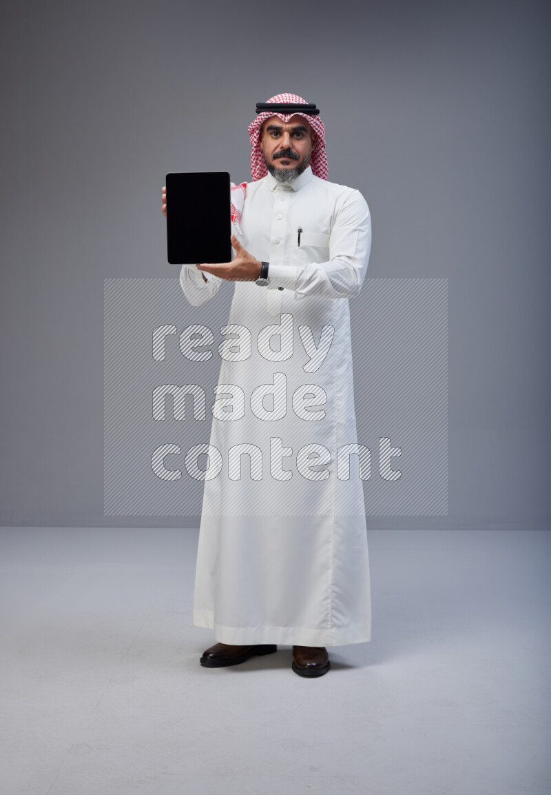 Saudi man Wearing Thob and red Shomag standing showing tablet to camera on Gray background
