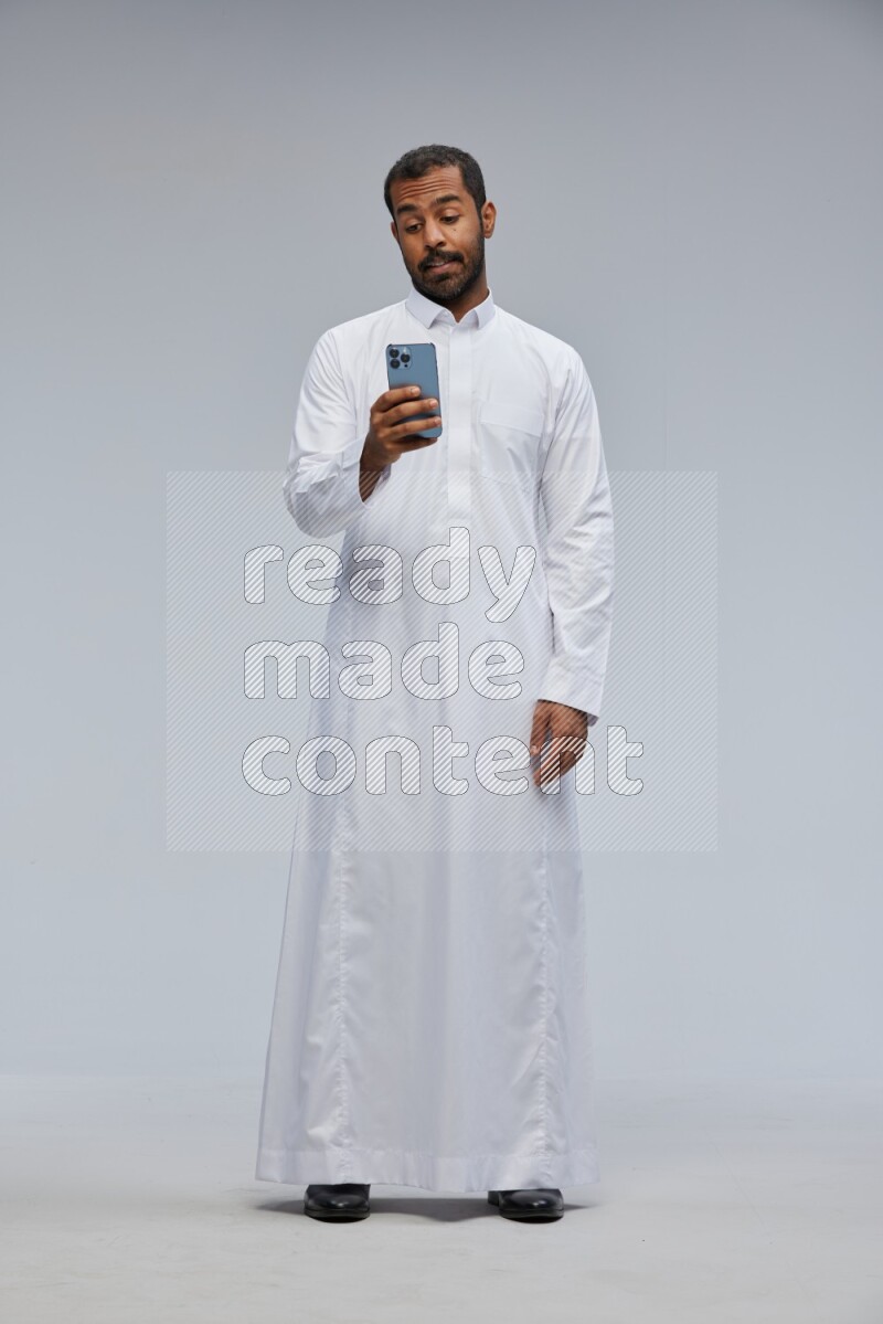 Saudi man wearing Thob standing texting on phone on Gray background