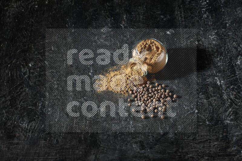 A flipped glass spice jar full of allspice powder and powder spilled out of it with whole balls on a textured black flooring