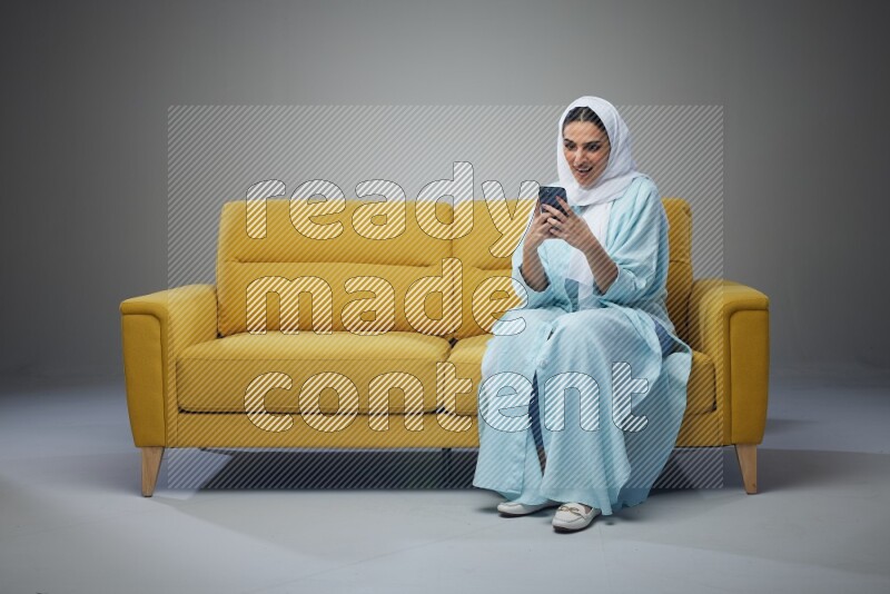 A Saudi woman wearing a light blue Abaya and white head scarf sitting on a yellow sofa and using her phone eye level on a grey background
