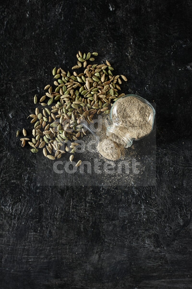 وعاء زجاجي للتوابل مقلوب وملعقة معدنية ممتلئة ببودرة الهيل وحبوب الهيل متناثرة على خلفية سوداء