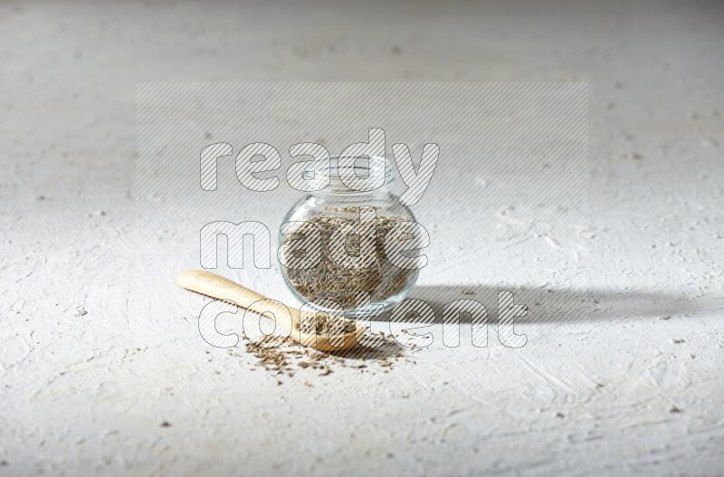 A glass spice jar and wooden spoon full of cumin seeds on textured white flooring