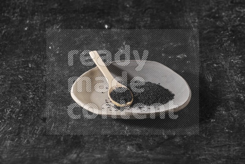 A multicolored pottery plate full of black seeds and wooden spoon full of seeds on a textured black flooring